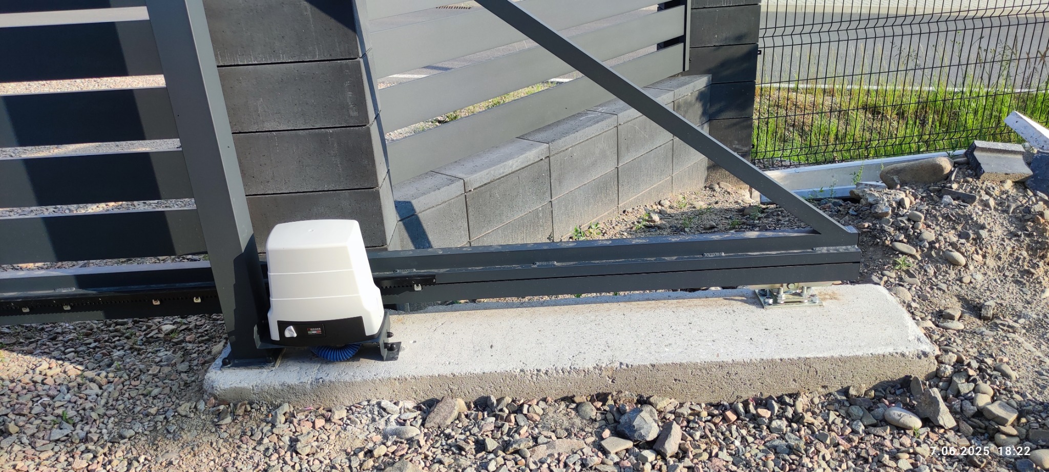 Szara brama przesuwna z automatyką, widok z bliska na zamontowany napęd na betonowej podstawie. Tło: ogrodzenie panelowe i gruz.