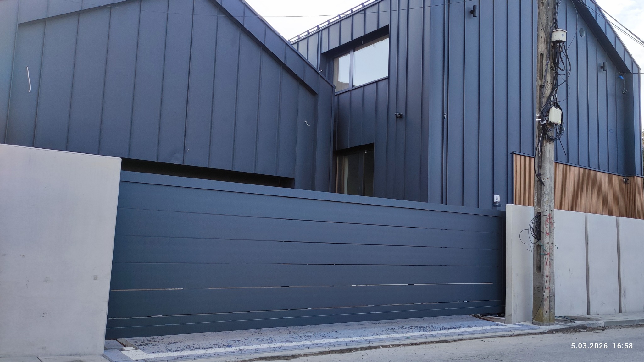 Nowoczesna, przesuwna brama wjazdowa w kolorze grafitowym, z poziomymi panelami, na tle domu o ciemnej elewacji i betonowego ogrodzenia. Widoczny słup energetyczny.