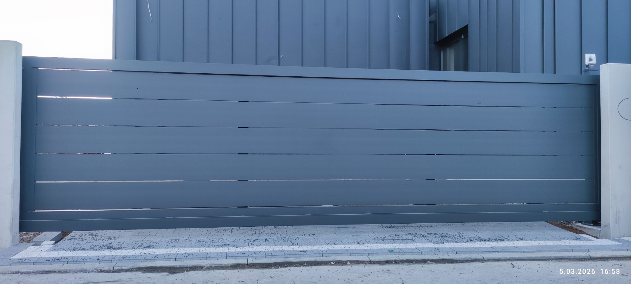 Nowoczesna, przesuwna brama wjazdowa w odcieniach szarości, z poziomymi panelami i widocznymi szczelinami, na tle elewacji budynku o podobnej kolorystyce.