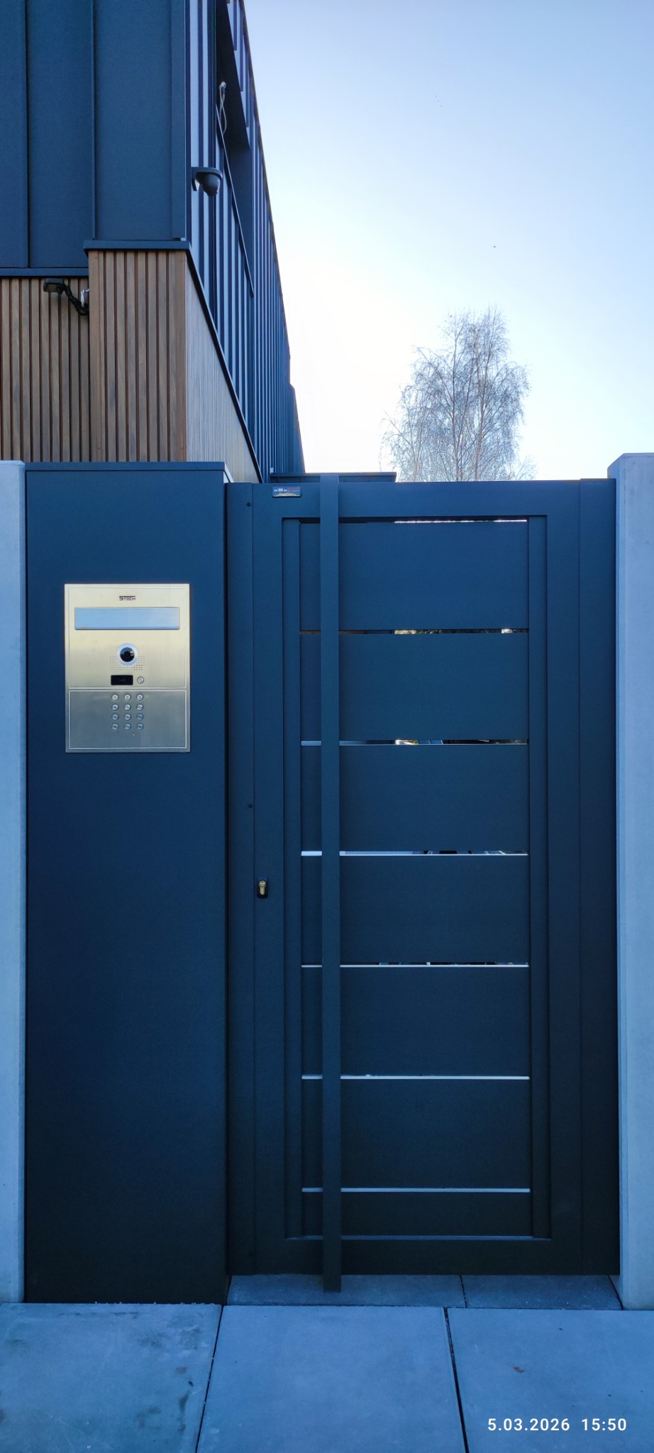 Nowoczesna, grafitowa brama wjazdowa z panelem domofonowym i poziomymi szczelinami, wkomponowana w architekturę domu z elementami drewna i betonu.
