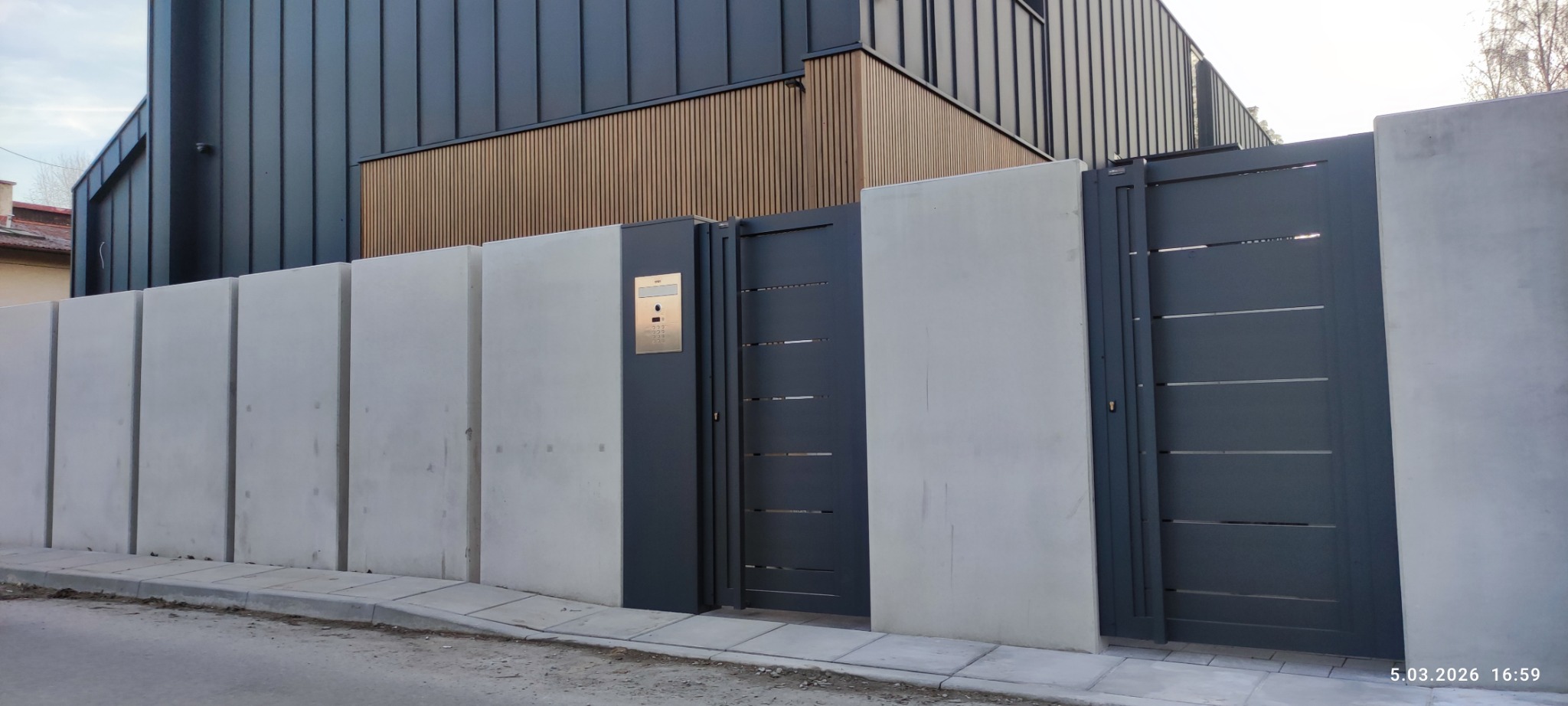 Nowoczesna, grafitowa brama wjazdowa z poziomymi szczeblami, wkomponowana w betonowy mur. Na słupku bramy widoczny panel domofonu.