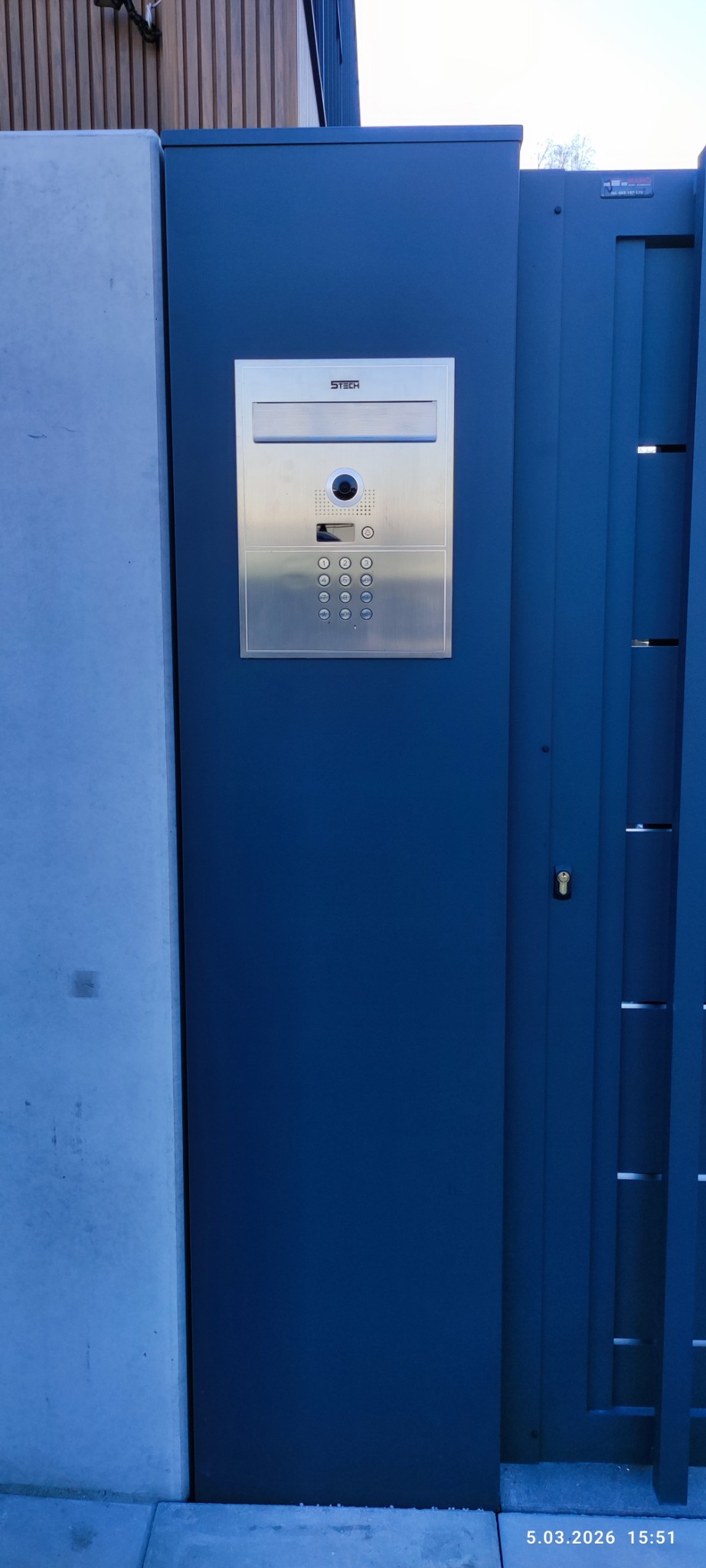 Nowoczesna brama wjazdowa z domofonem Stetech w Wiśniowej. Metalowa konstrukcja w odcieniach szarości i niebieskiego. Minimalistyczny design.