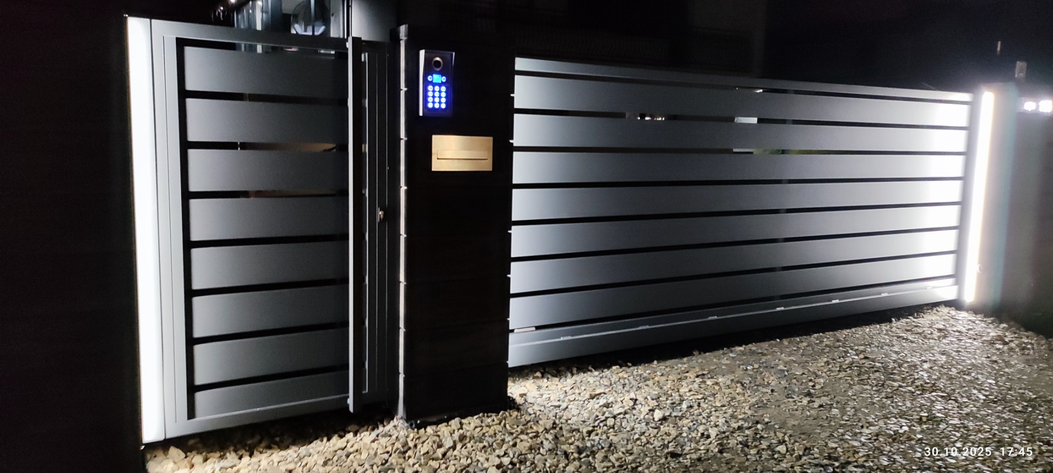 Nowoczesna, przesuwna brama wjazdowa z podświetleniem LED, wideodomofonem i skrzynką na listy. Szara kolorystyka, poziome panele, oświetlenie nocne.