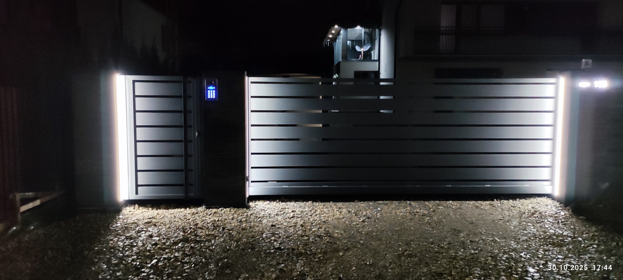 Nowoczesna brama wjazdowa z poziomymi panelami i oświetleniem LED, częściowo otwarta, z panelem sterowania numerycznego. Nocne ujęcie.