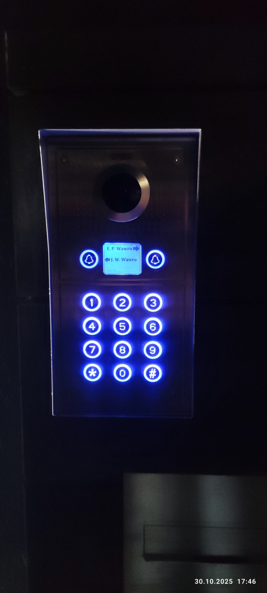 Panel domofonu z podświetlanymi przyciskami i wyświetlaczem nazwisk, zamontowany na ciemnej powierzchni. Widoczne cyfry i symbole.