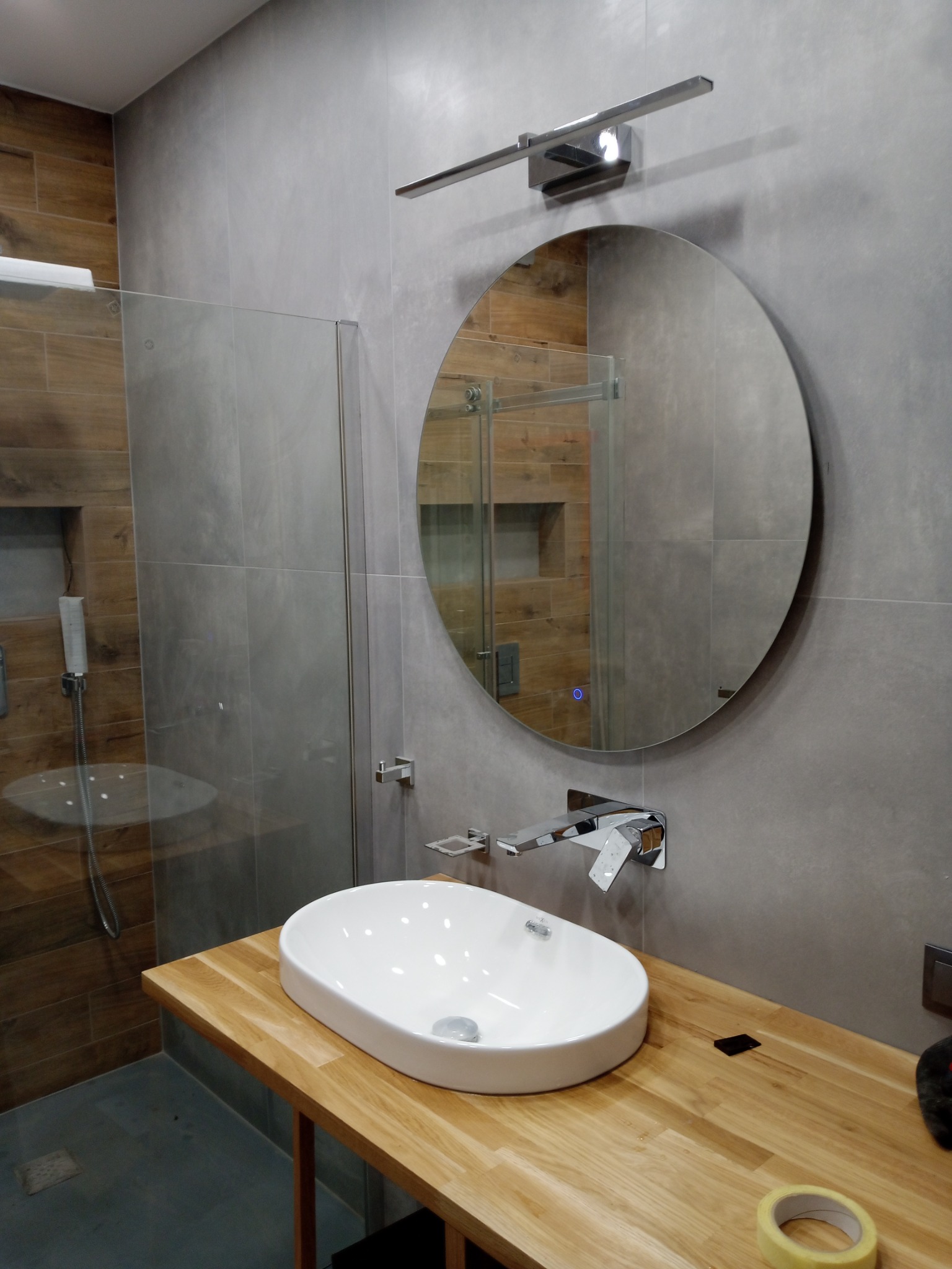 Nowoczesna łazienka z owalną umywalką na drewnianym blacie, okrągłym lustrem z podświetleniem i kabiną prysznicową z szarymi płytkami imitującymi beton.