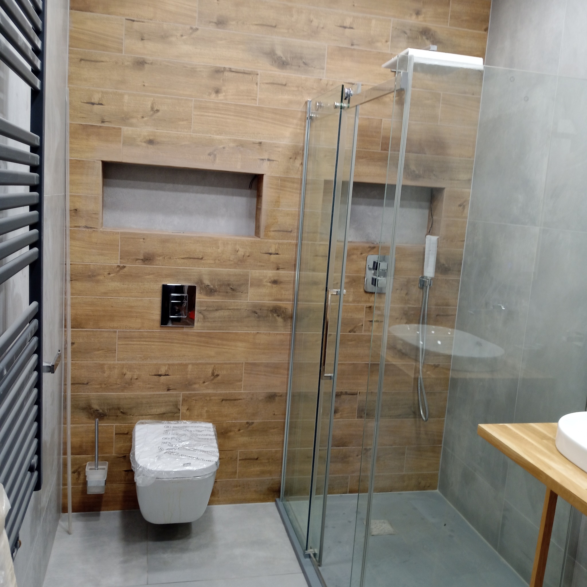 Nowoczesna łazienka z drewnopodobnymi płytkami, kabiną prysznicową ze szkła i wiszącą toaletą. Grzejnik drabinkowy po lewej stronie. Wnęki w ścianie.