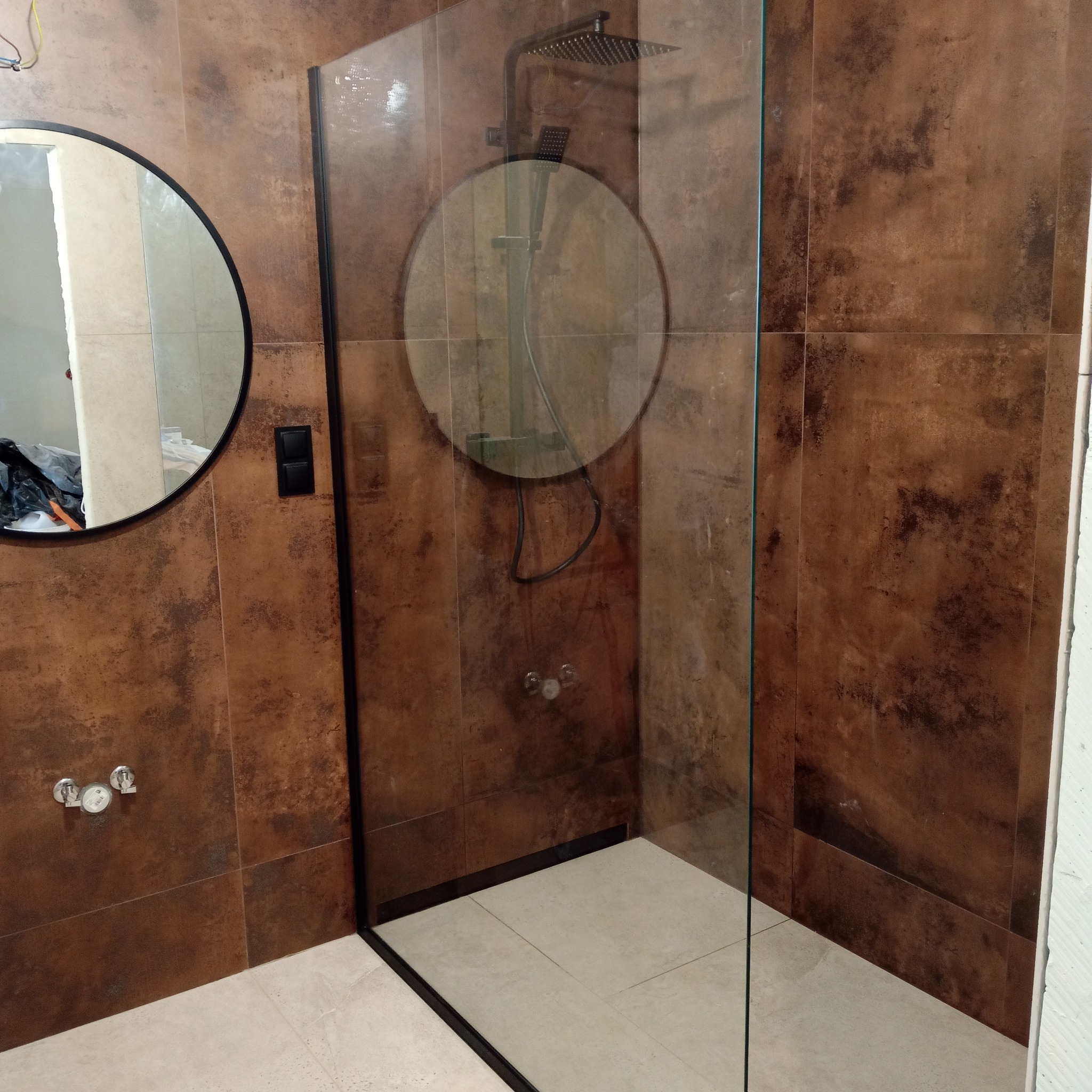 Nowoczesna kabina prysznicowa z czarną ramą i deszczownicą, okrągłe lustro na ścianie z brązowych płytek. Minimalistyczny design łazienki.
