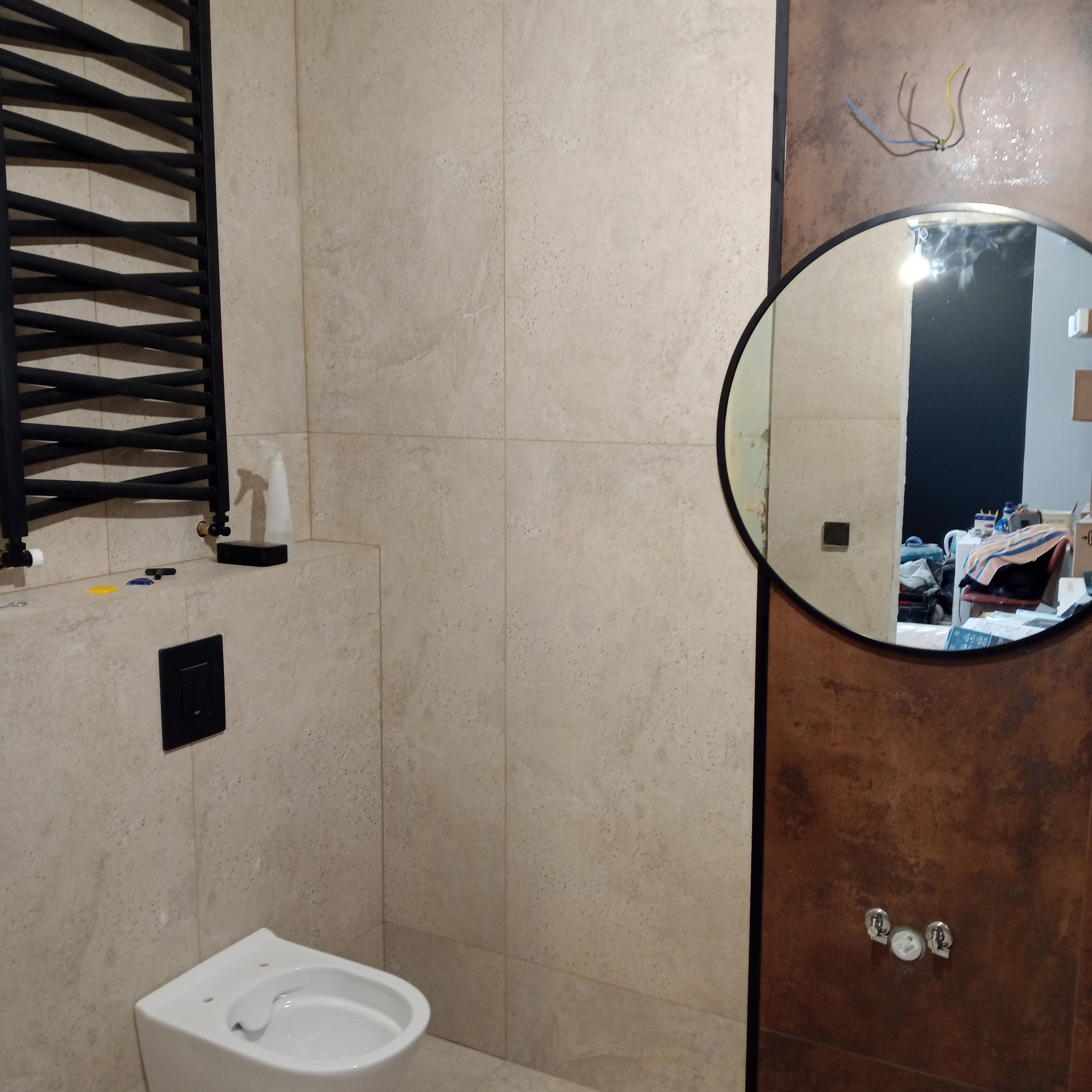 Wykończona łazienka z beżowymi płytkami, grzejnikiem drabinkowym i okrągłym lustrem, w którym odbija się wnętrze. Widoczne wystające kable.