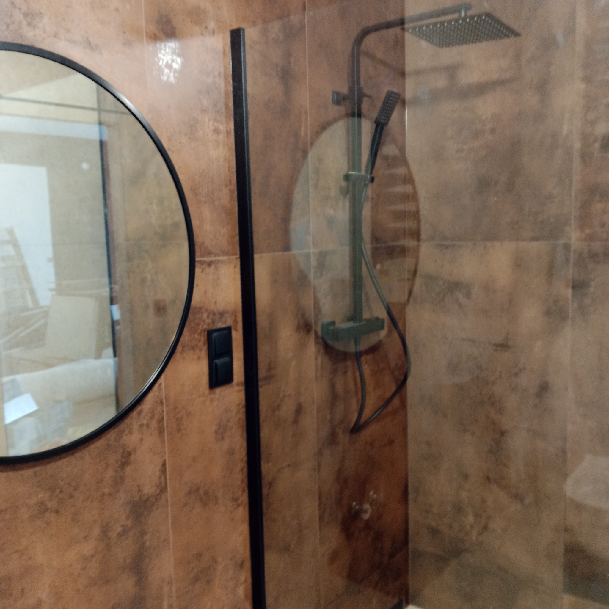 Nowoczesna kabina prysznicowa z brązowymi płytkami imitującymi kamień, okrągłe lustro z czarną ramą i czarny panel sterowania.