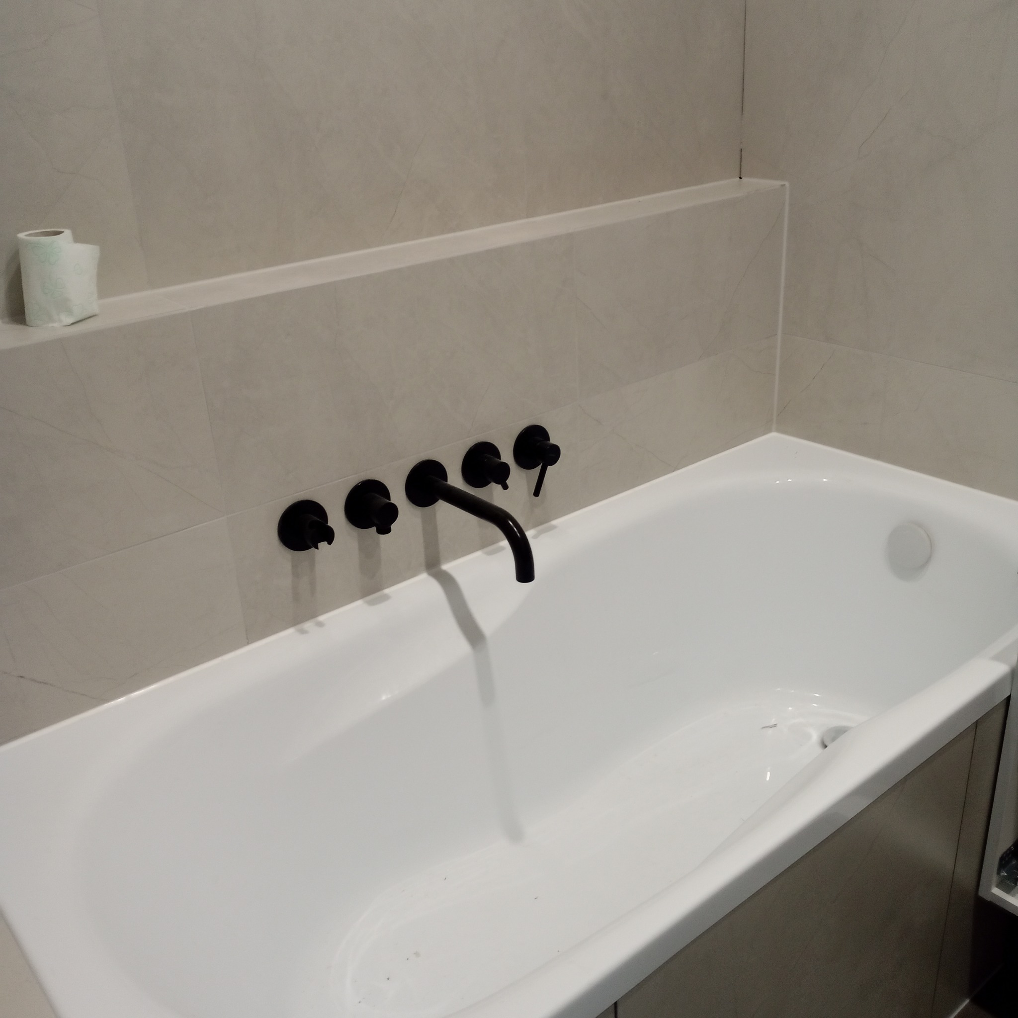 Biała wanna z czarną armaturą na tle beżowych płytek. Minimalistyczny design łazienki z rolką papieru toaletowego w rogu.