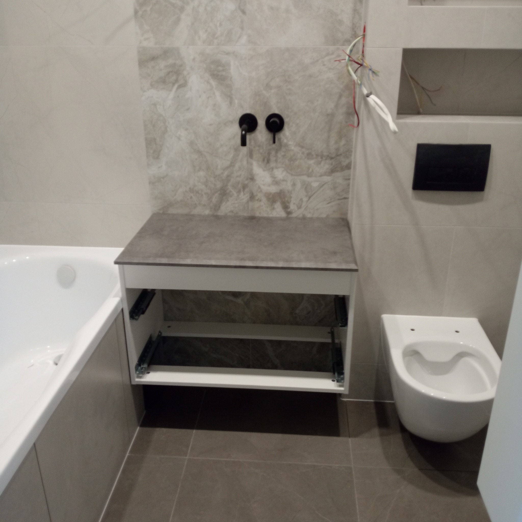 Nowoczesna łazienka w szarościach: wisząca toaleta, wanna i szafka pod umywalkę z czarną armaturą. Widoczne płytki imitujące marmur i wystające przewody.