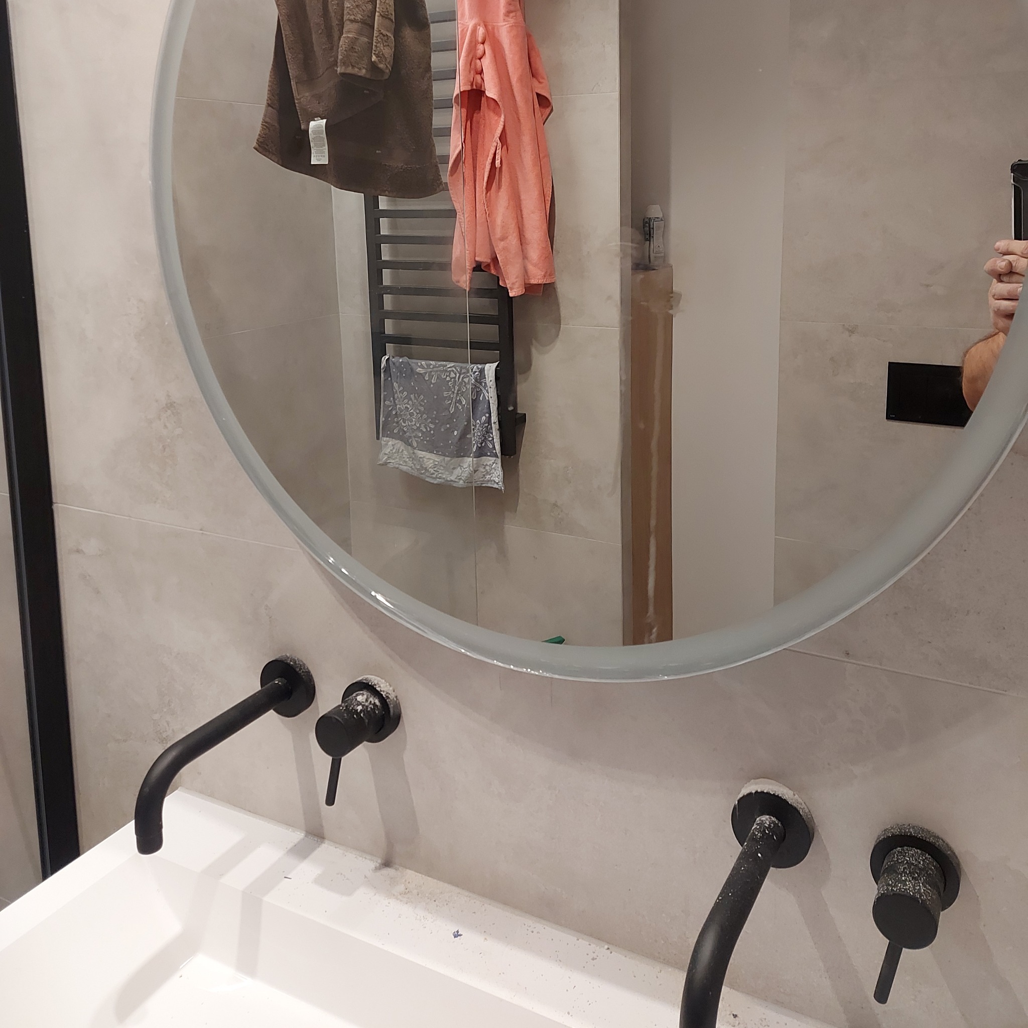 Wykończona łazienka z okrągłym lustrem, czarną armaturą i beżowymi płytkami. Widoczne detale wykończeniowe i odbicie fragmentu osoby robiącej zdjęcie.