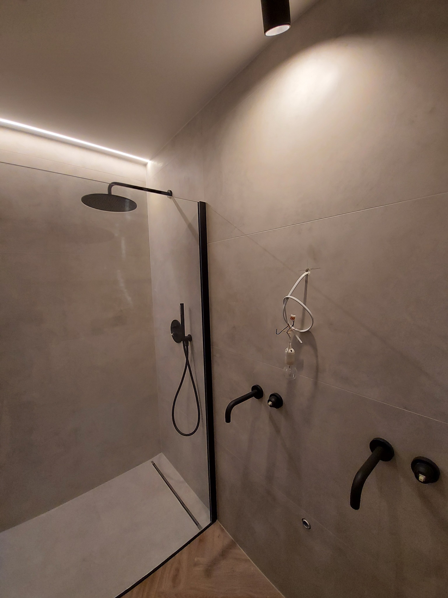 Nowoczesna kabina prysznicowa walk-in z czarnymi elementami armatury i odpływem liniowym, oświetlona ledowym paskiem i punktowym światłem. Widoczne płytki w odcieniach szarości.