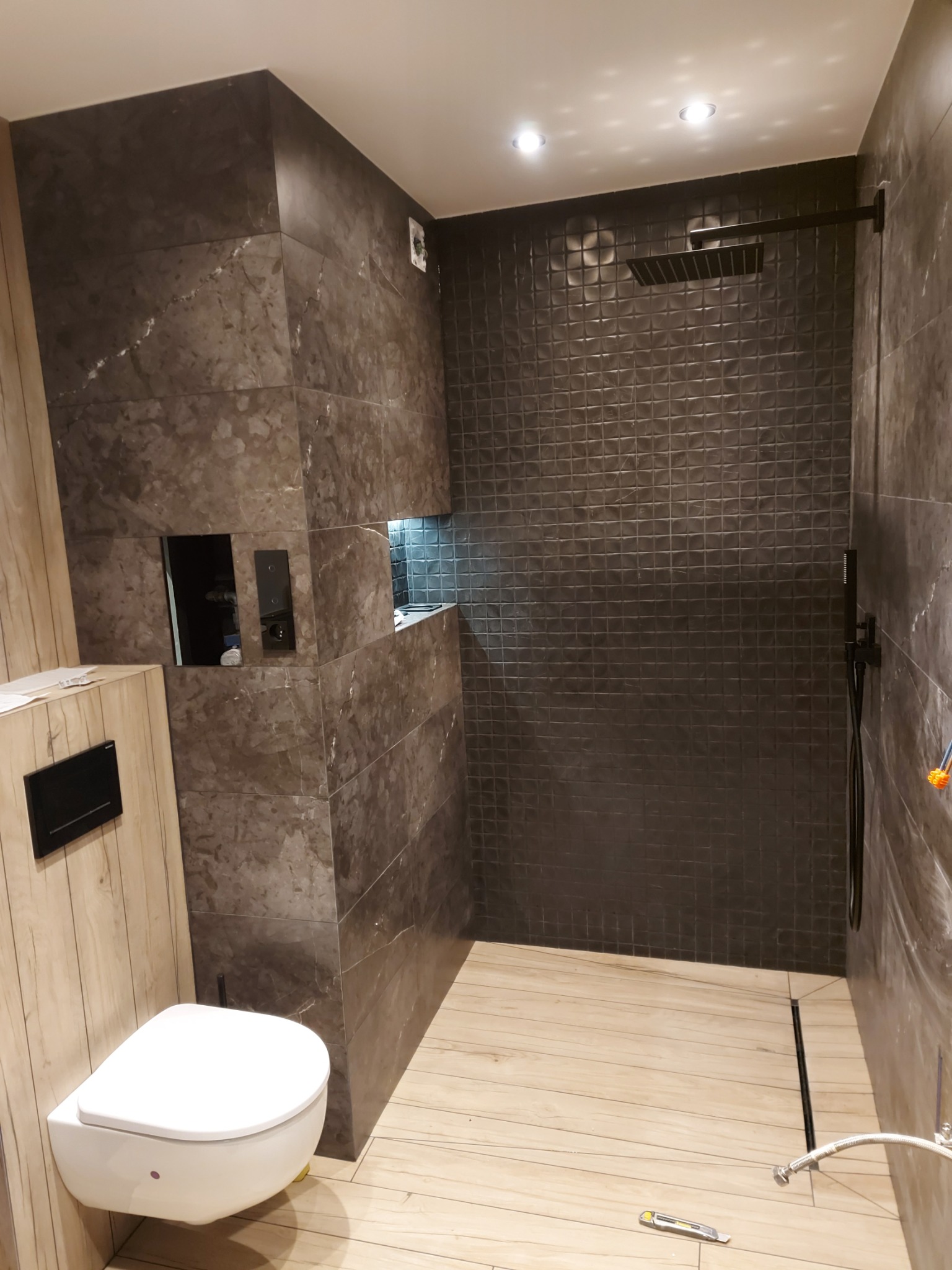 Nowoczesna łazienka z kabiną typu walk-in, wykończona płytkami imitującymi marmur i drewno. Widoczna toaleta podwieszana i czarna armatura.