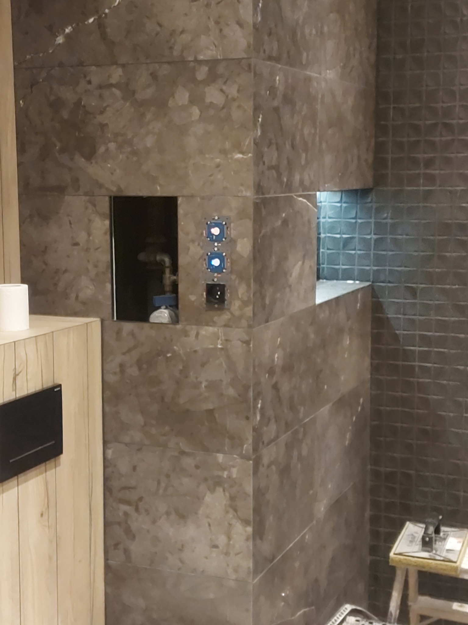 Wykończona ściana w łazience z brązowych płytek imitujących kamień, z wnęką i widoczną instalacją hydrauliczną oraz fragmentem mozaiki na sąsiedniej ścianie.