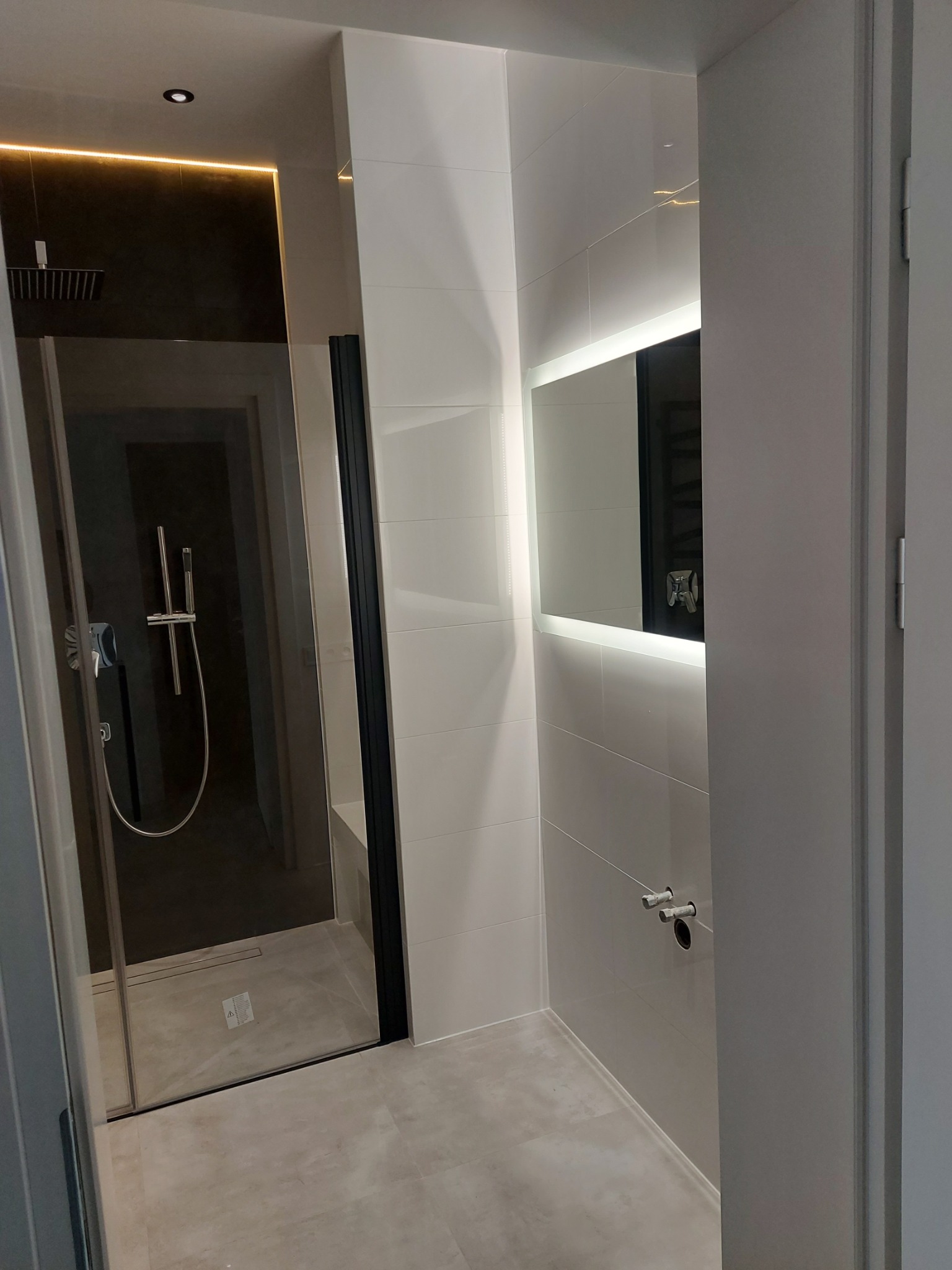 Nowoczesna łazienka z kabiną prysznicową typu walk-in, podświetlanym lustrem i jasnymi płytkami. Minimalistyczny design z akcentami światła.