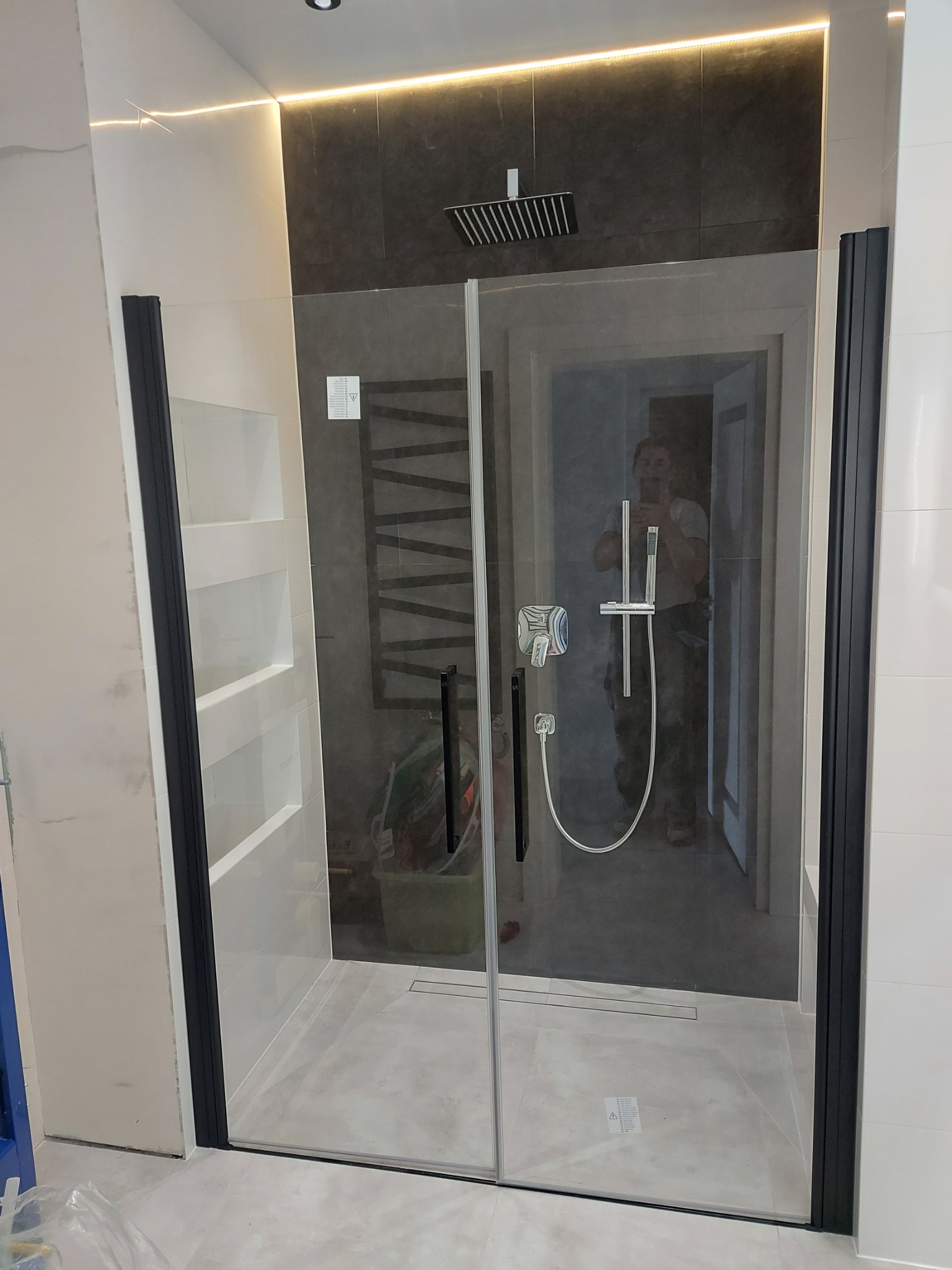 Nowoczesna kabina prysznicowa z czarnymi profilami i deszczownicą, wykończona w jasnych płytkach z akcentem ciemnej ściany i podświetleniem LED. Widoczny odpływ liniowy.