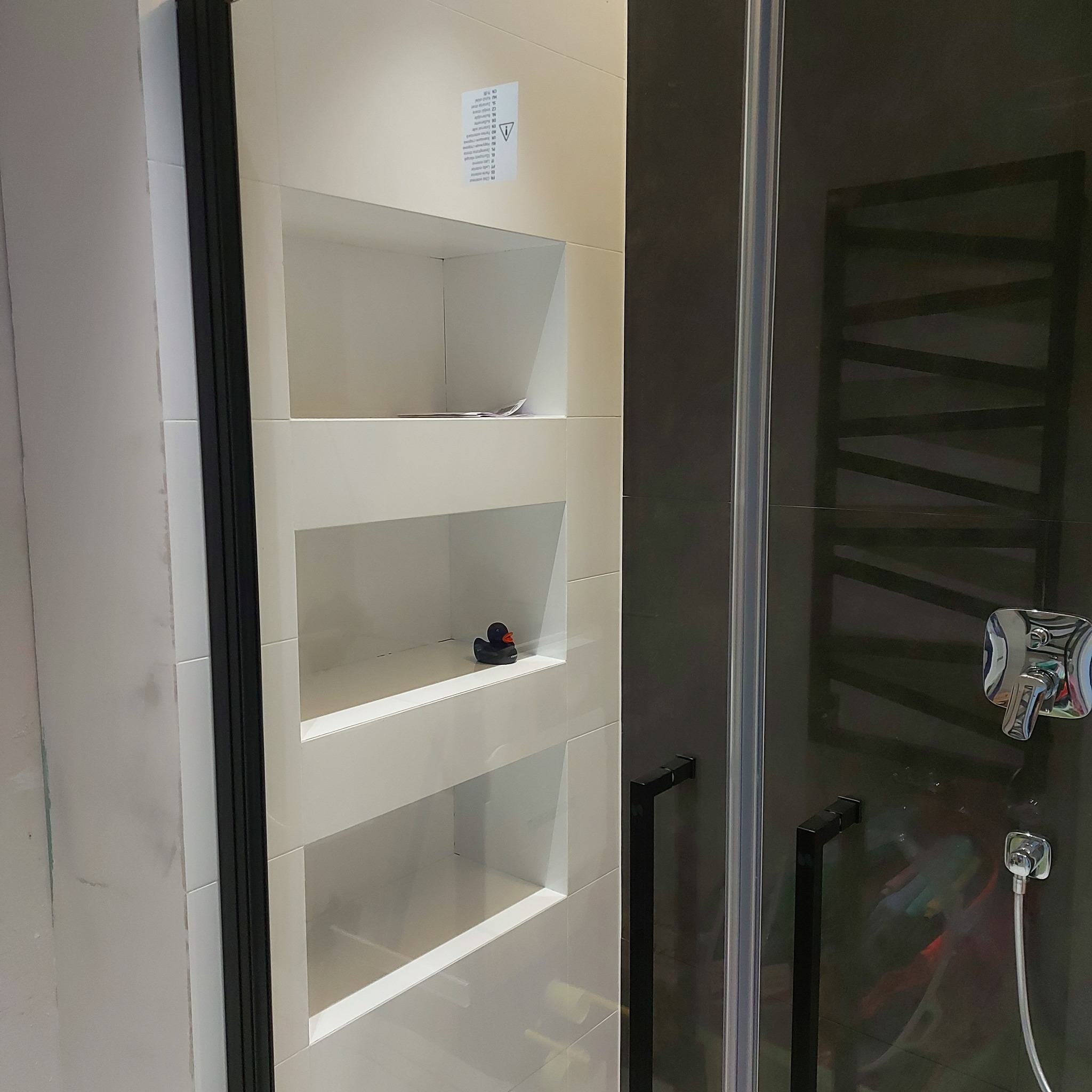 Biała, kafelkowana wnęka z trzema półkami w łazience, z czarną gumową kaczką na środkowej półce, obok kabina prysznicowa z czarnymi elementami.