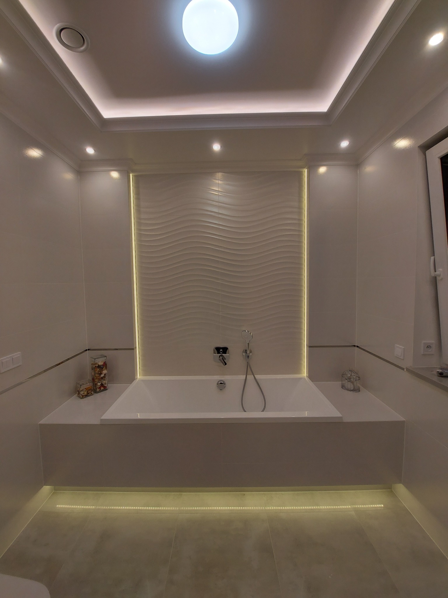 Nowoczesna łazienka z wanną, podświetleniem LED na suficie i pod wanną. Na ścianie dekoracyjna płytka z falistym wzorem. Minimalistyczny design.