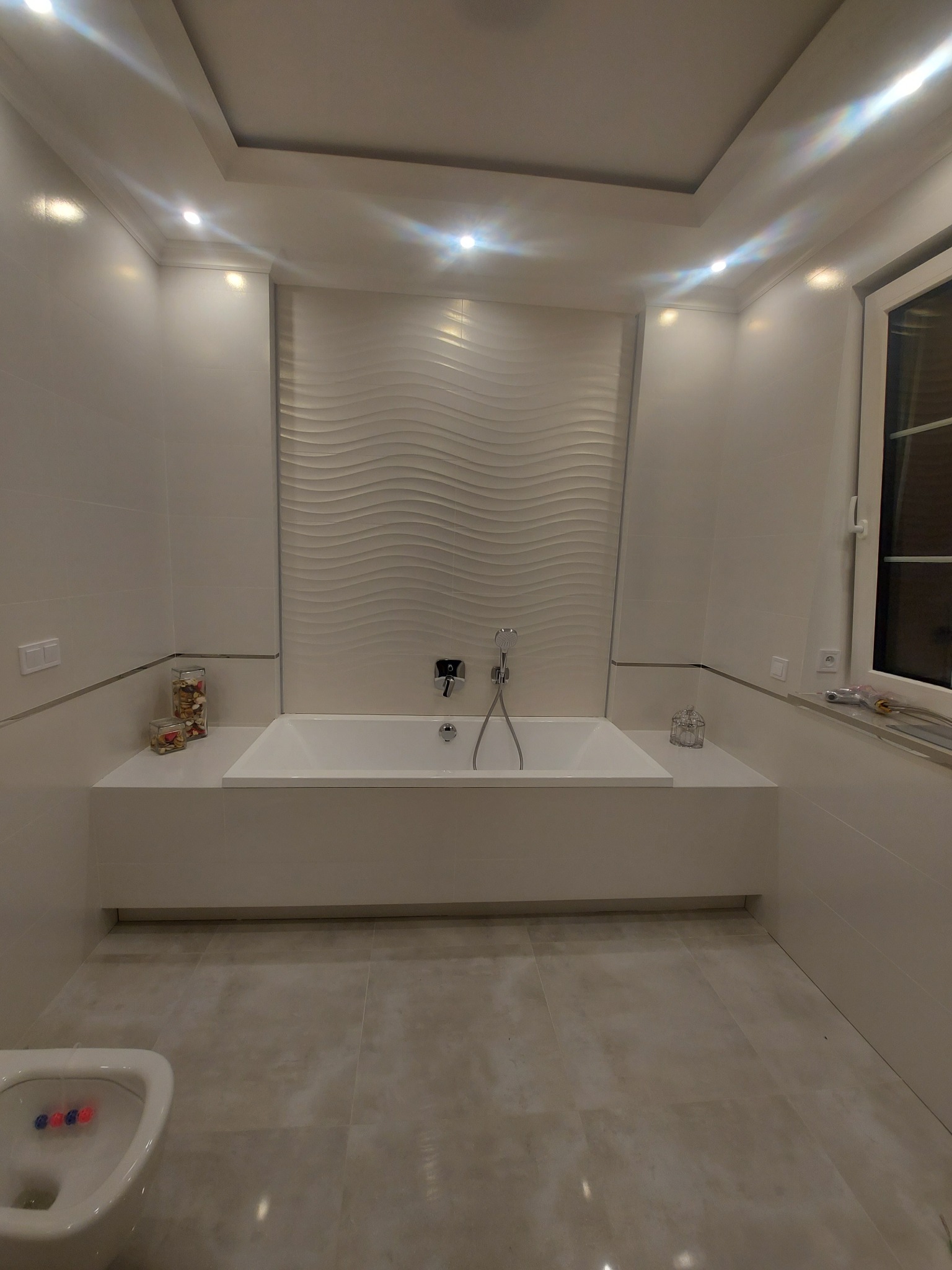 Elegancka łazienka w jasnych barwach z wanną obudowaną płytkami, dekoracyjną ścianą z motywem fal i subtelnym oświetleniem punktowym.