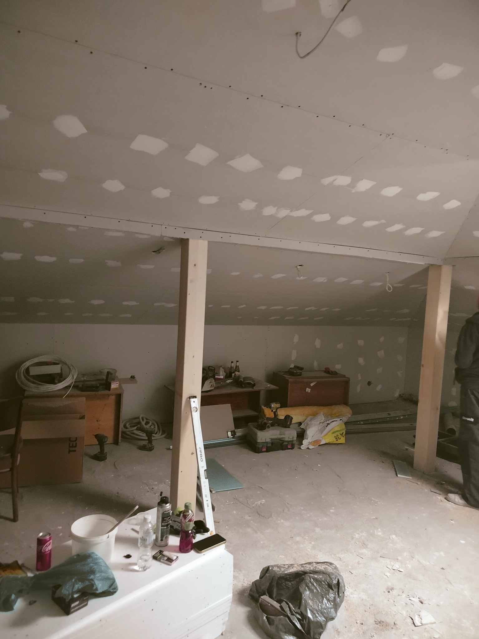Poddasze w trakcie wykańczania płytami GK, widoczne drewniane słupy, narzędzia i materiały budowlane. Ślady szpachlowania na łączeniach płyt. Ujęcie z dołu, perspektywa wnętrza.