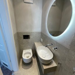 TIP-TOP GLAZURNIK Marcin Kapuściński - Nowoczesna łazienka w szarościach: podwieszana toaleta, owalna umywalka na kamiennym blacie i podświetlane lustro. Minimalistyczny design z dbałością o detale.