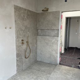 TIP-TOP GLAZURNIK Marcin Kapuściński - Wykończona kabina prysznicowa z szarymi płytkami imitującymi kamień, złotą armaturą i odpływem liniowym. Widoczne detale wykończenia i surowy stan ścian.