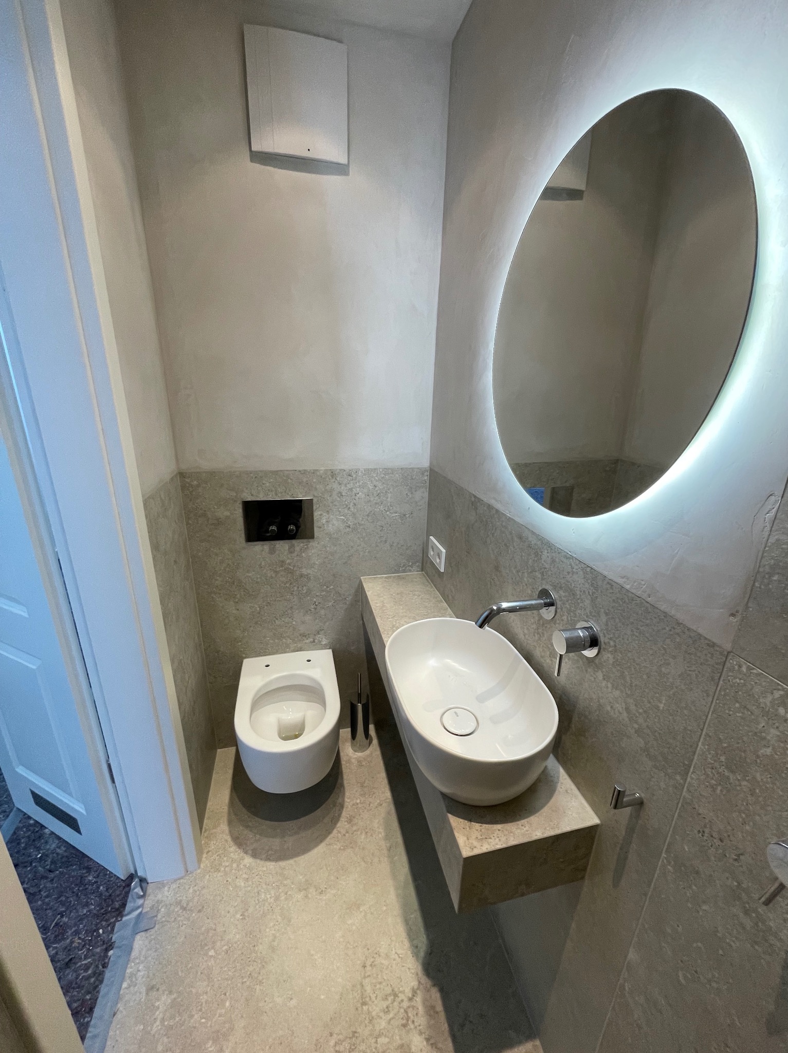 Nowoczesna łazienka w szarościach: podwieszana toaleta, owalna umywalka na kamiennym blacie i podświetlane lustro. Minimalistyczny design z dbałością o detale.