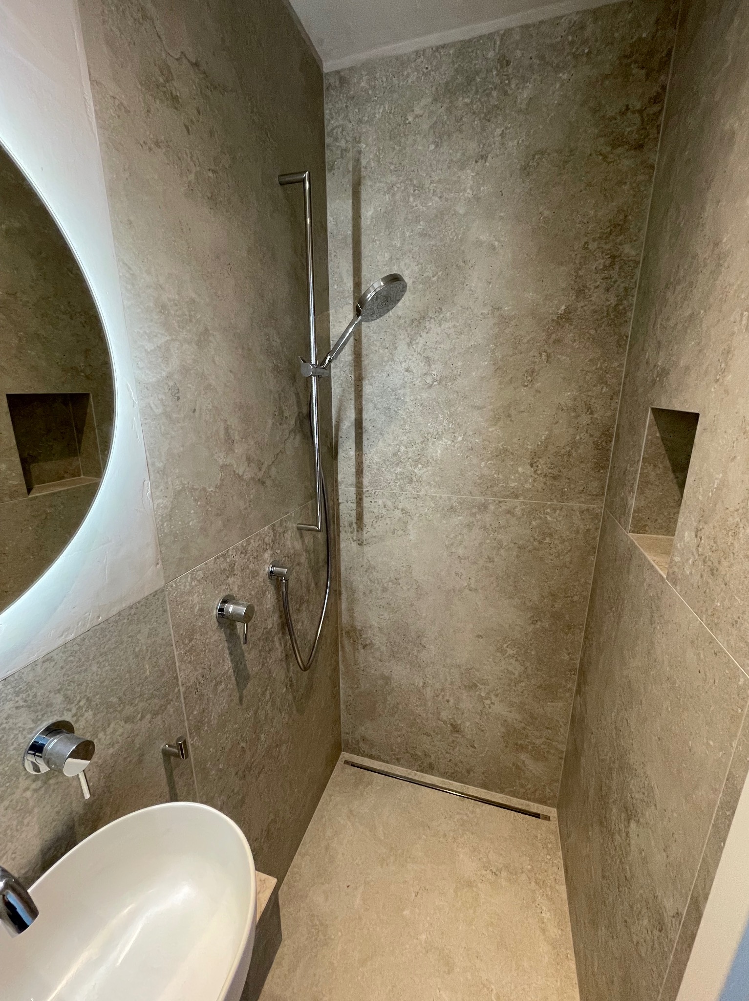 Beżowa łazienka z owalną umywalką, chromowaną armaturą i kabiną prysznicową typu walk-in. W ścianie wnęka. Minimalistyczny design, neutralna kolorystyka.