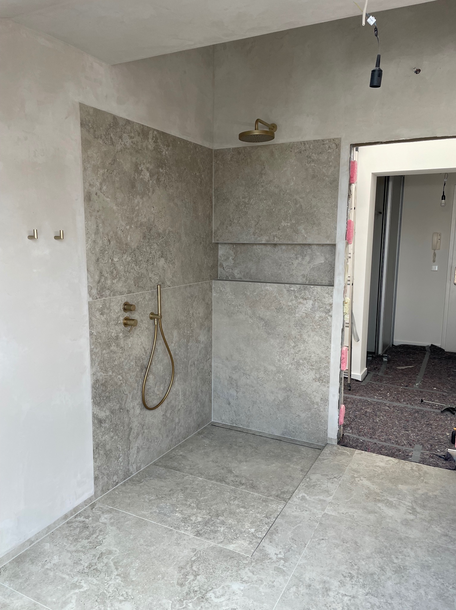 Wykończona kabina prysznicowa z szarymi płytkami imitującymi kamień, złotą armaturą i odpływem liniowym. Widoczne detale wykończenia i surowy stan ścian.