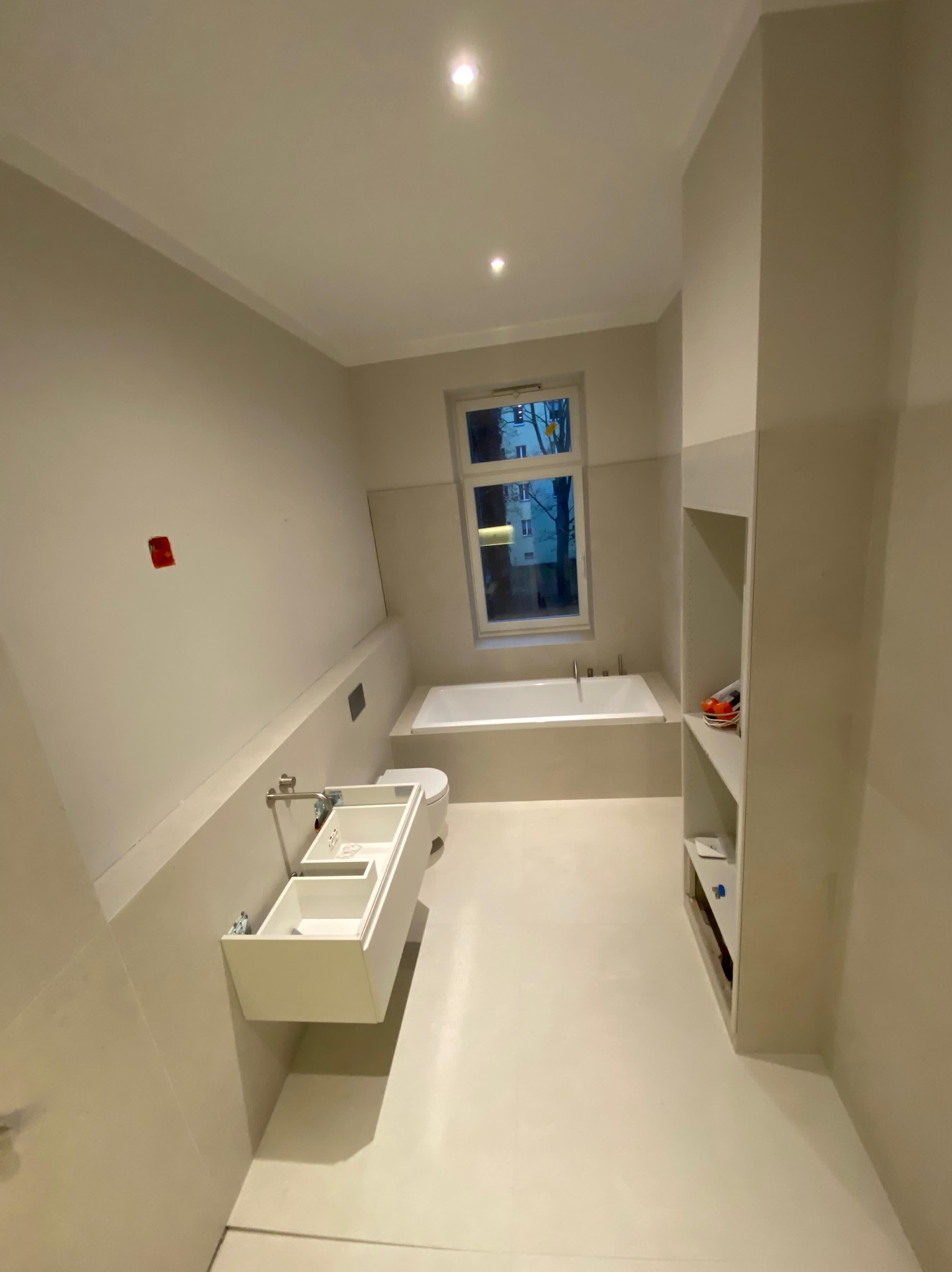 Jasna łazienka w trakcie wykańczania: biała wanna pod oknem, wisząca szafka umywalkowa bez frontów, półki w ścianie. Widoczne niedokończone prace.