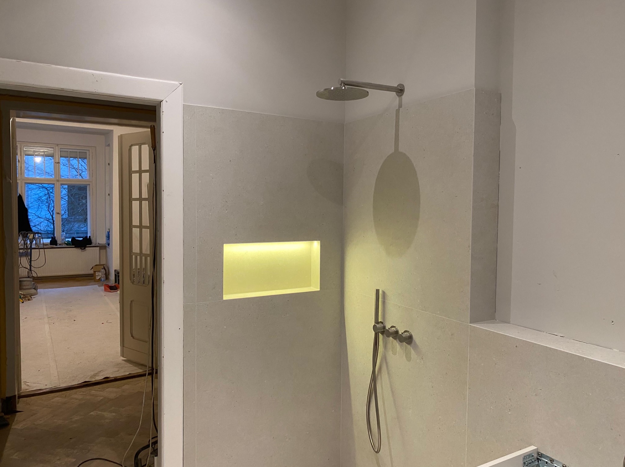 Wykończona strefa prysznicowa z deszczownicą i wnęką z podświetleniem w łazience. Widok na remontowane pomieszczenie przez otwarte drzwi.