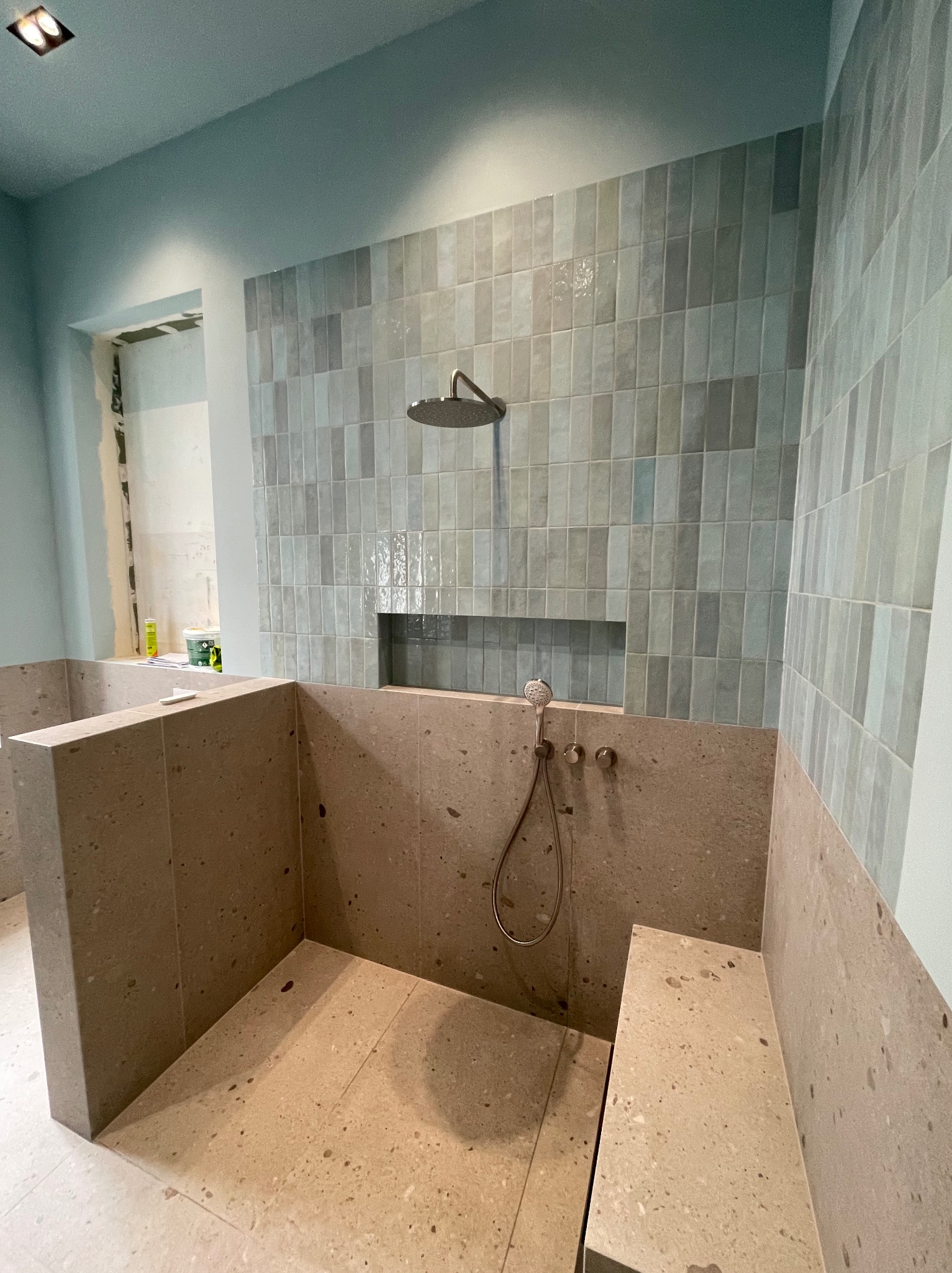 Wykończona kabina prysznicowa z beżową mozaiką na podłodze i ścianach, oraz z szaro-niebieskimi płytkami na ścianie z prysznicem. Widoczna słuchawka i deszczownica.