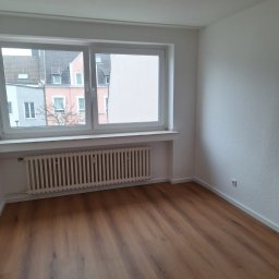 Kacper Gniewowski Wykończenia Wnętrz - Jasne wnętrze pokoju po remoncie: drewnopodobna podłoga, białe ściany i grzejnik pod oknem z widokiem na budynki. Minimalistyczny design, świeżo po wykończeniu.