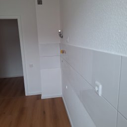 Kacper Gniewowski Wykończenia Wnętrz - Białe, błyszczące płytki na ścianie w rogu pomieszczenia, widoczne gniazdka elektryczne i wieszak. Podłoga wyłożona panelami imitującymi drewno. Widok na otwarte drzwi.