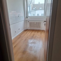 Kacper Gniewowski Wykończenia Wnętrz - Jasne drewniane panele w pustym pokoju z białymi ścianami i grzejnikiem pod oknem. Widok zza otwartych drzwi, z fragmentem białych płytek na ścianie.