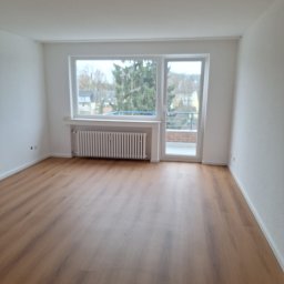 Kacper Gniewowski Wykończenia Wnętrz - Pusty pokój z panelami podłogowymi, białymi ścianami i grzejnikiem pod oknem balkonowym z widokiem na zieleń. Świeżo wykończone wnętrze.