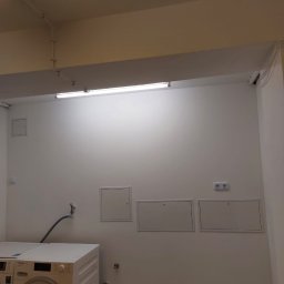 Białe, odświeżone pomieszczenie gospodarcze z pralką Miele, trzema rewizjami na ścianie i nowym oświetleniem. Ściany pomalowane na jednolity, jasny kolor.