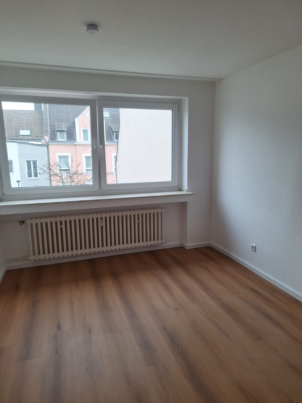 Jasne wnętrze pokoju po remoncie: drewnopodobna podłoga, białe ściany i grzejnik pod oknem z widokiem na budynki. Minimalistyczny design, świeżo po wykończeniu.