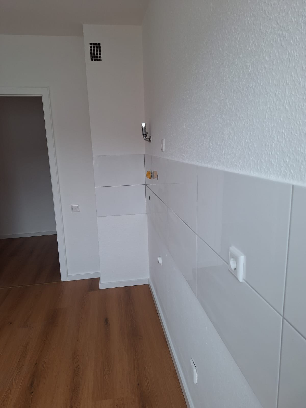 Białe, błyszczące płytki na ścianie w rogu pomieszczenia, widoczne gniazdka elektryczne i wieszak. Podłoga wyłożona panelami imitującymi drewno. Widok na otwarte drzwi.