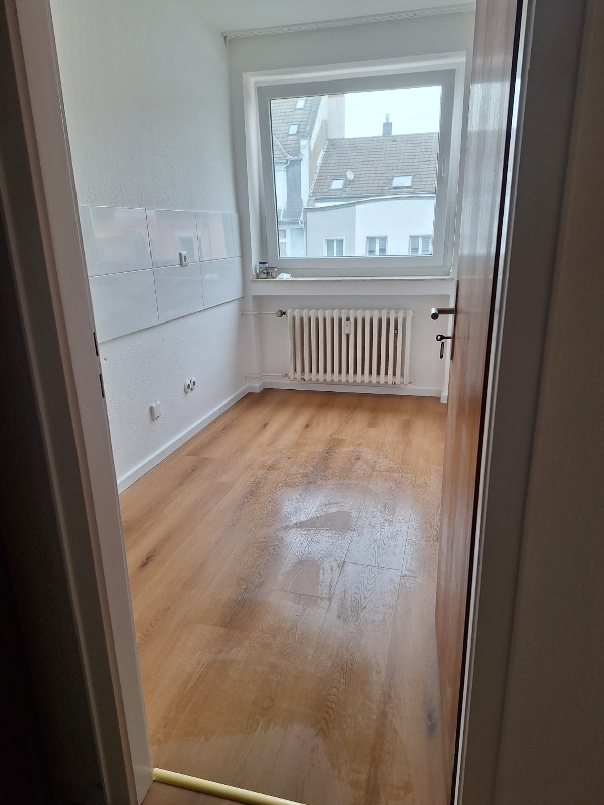 Jasne drewniane panele w pustym pokoju z białymi ścianami i grzejnikiem pod oknem. Widok zza otwartych drzwi, z fragmentem białych płytek na ścianie.