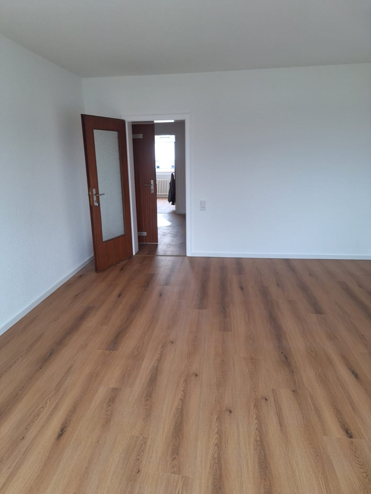 Jasne panele podłogowe w pustym pokoju z białymi ścianami i otwartymi drzwiami z brązowymi futrynami. Widok na sąsiednie pomieszczenie.