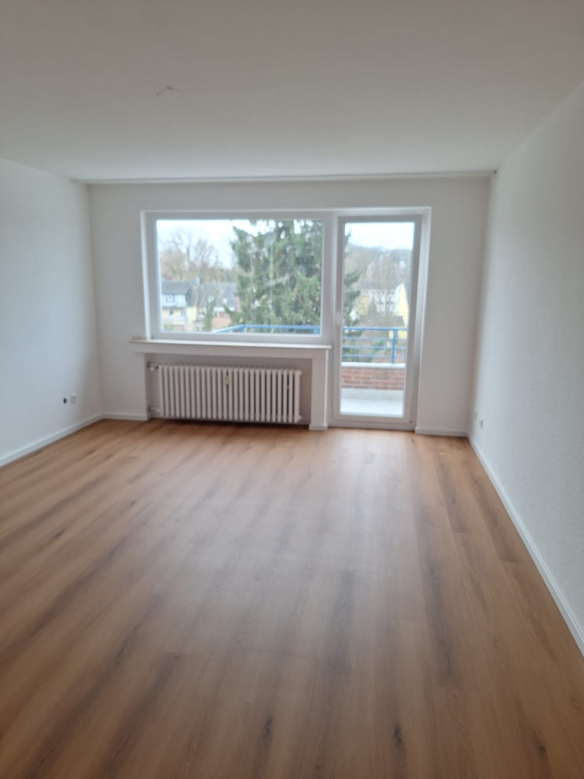 Pusty pokój z panelami podłogowymi, białymi ścianami i grzejnikiem pod oknem balkonowym z widokiem na zieleń. Świeżo wykończone wnętrze.