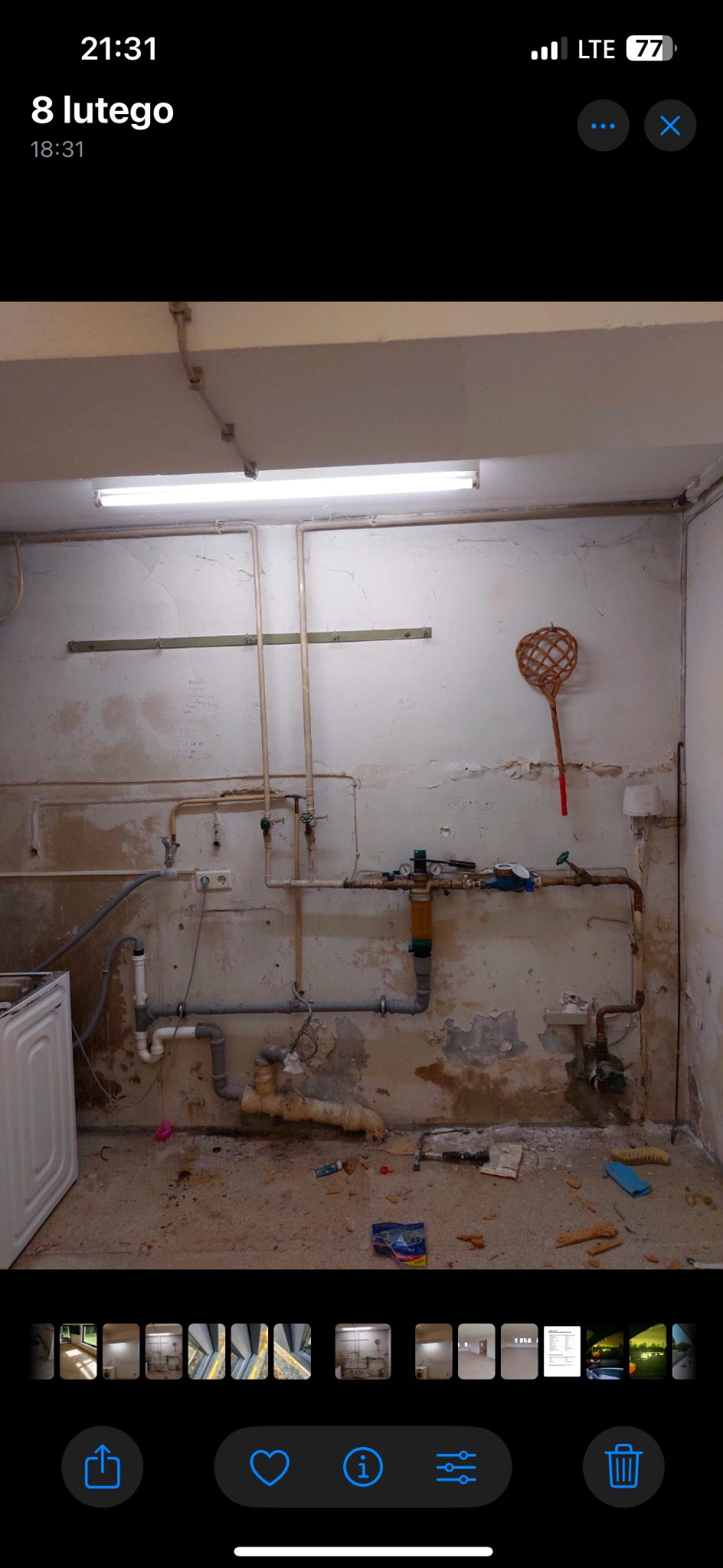 Zniszczone wnętrze pomieszczenia przed remontem: widoczne stare instalacje, odpadający tynk, zabrudzona podłoga i ściany. W rogu stoi pralka.