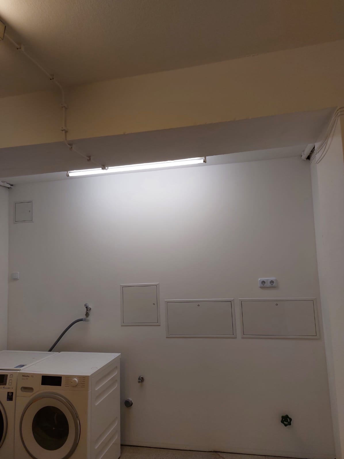 Białe, odświeżone pomieszczenie gospodarcze z pralką Miele, trzema rewizjami na ścianie i nowym oświetleniem. Ściany pomalowane na jednolity, jasny kolor.