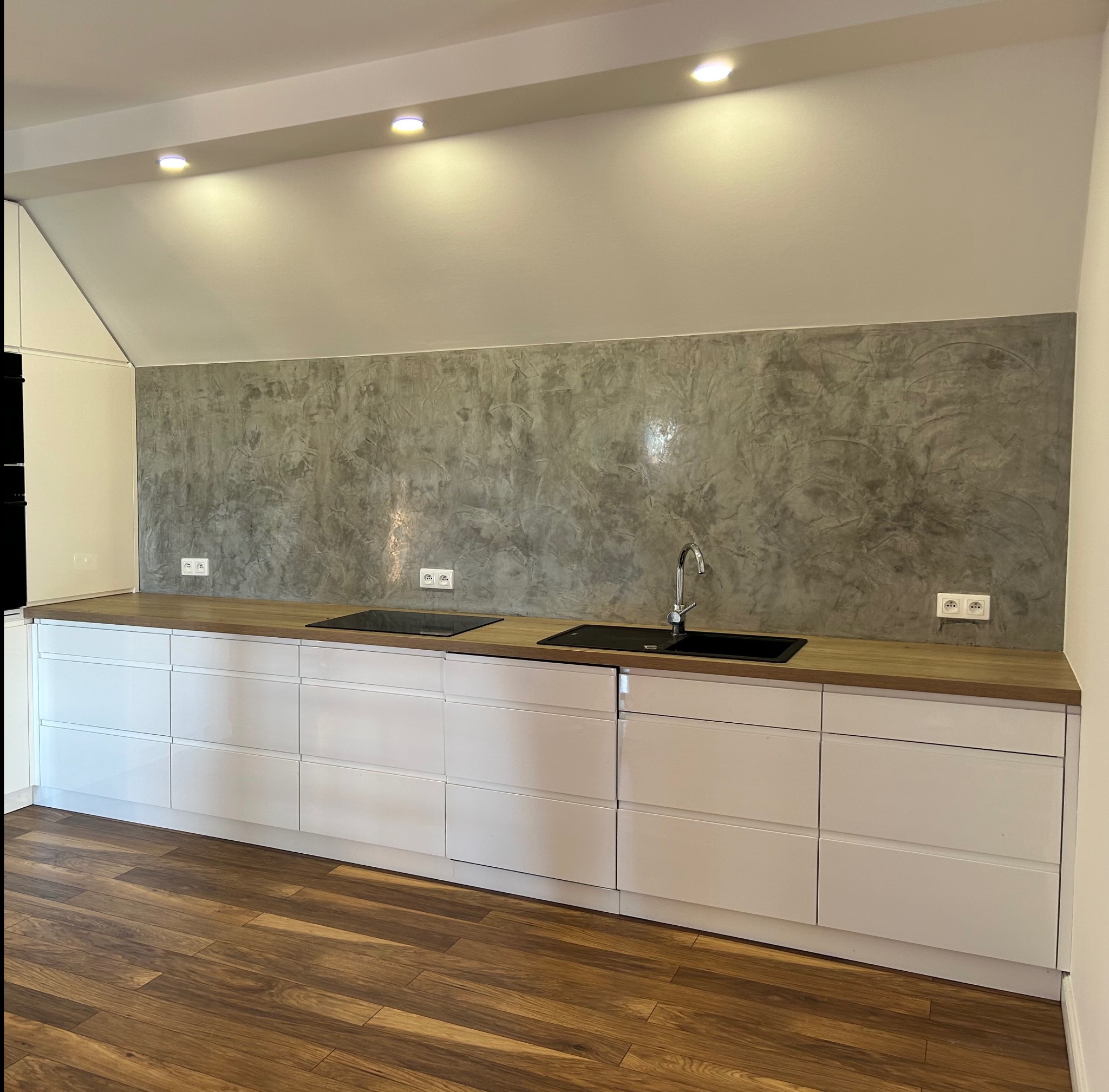 Kuchnia po remoncie: białe szafki, drewniany blat, ściana ze stiukiem w odcieniach szarości. Widoczne gniazdka elektryczne i płyta indukcyjna. Podłoga z paneli imitujących drewno.