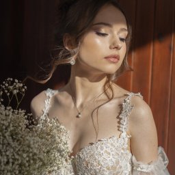 photovu - Panna młoda w koronkowej sukni, z bukietem gipsówki, w ciepłym świetle na tle drewnianej ściany. Subtelny makijaż i delikatna biżuteria podkreślają uroczysty charakter.