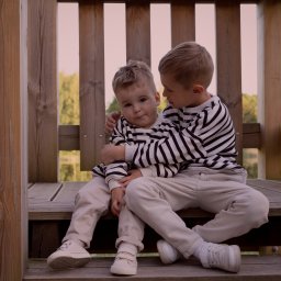 photovu - Dwóch chłopców w pasiaste bluzy siedzi na drewnianych schodach konstrukcji. Starszy obejmuje młodszego. Tło stanowią drewniane elementy i zieleń.