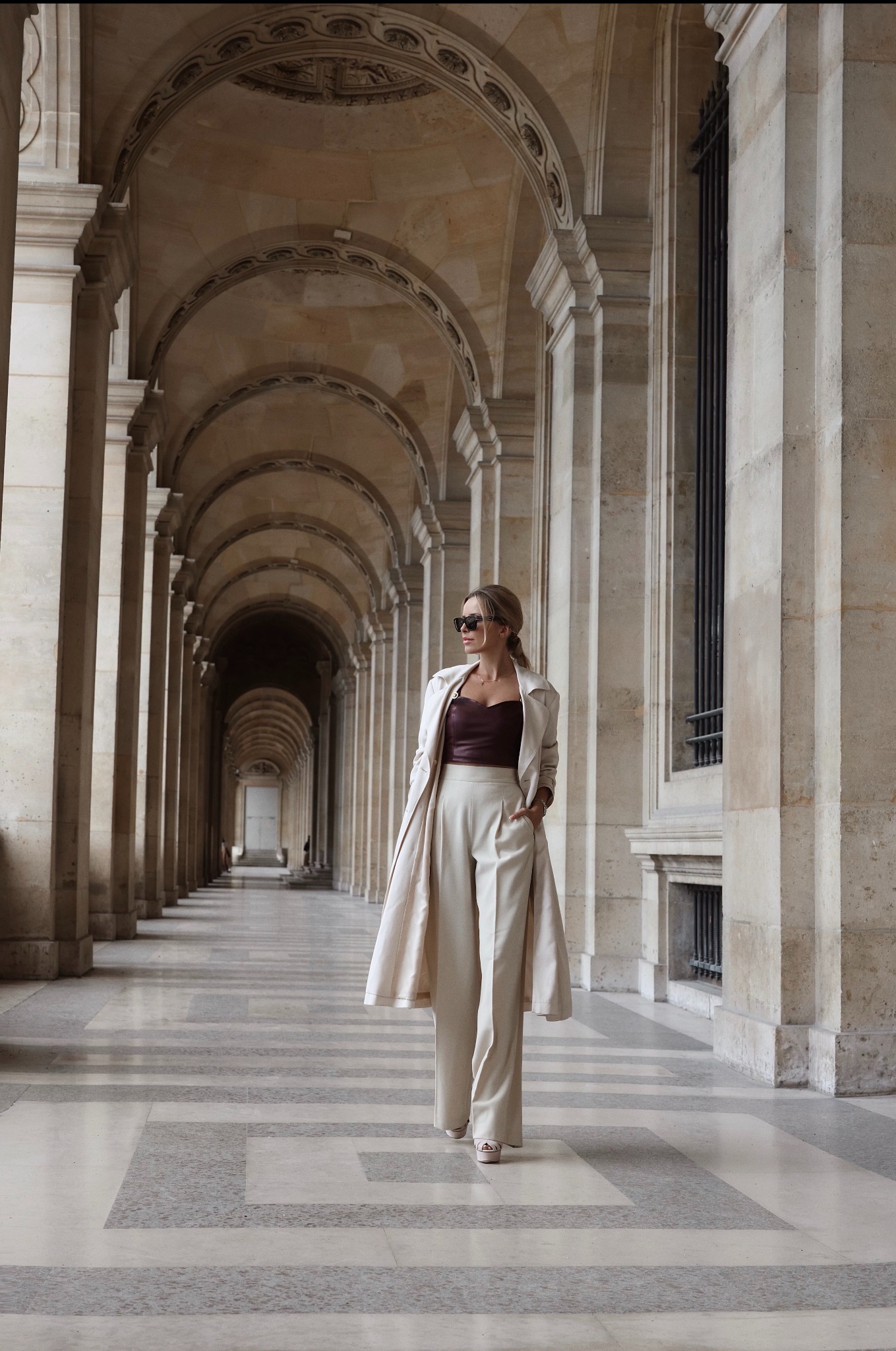 Kobieta w eleganckim, beżowym garniturze i okularach przeciwsłonecznych spaceruje po korytarzu z kolumnami i łukami. Stonowana kolorystyka, symetryczny kadr.